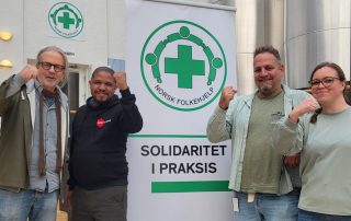 Arne Presterud, Thapelo Mohapi, Frank Toks og Naemy Trandum Aasen står foran en plakat med Norsk Folkehjelps logo og teksten "Solidaritet i praksis," mens de løfter knyttede never som symbol på solidaritet.