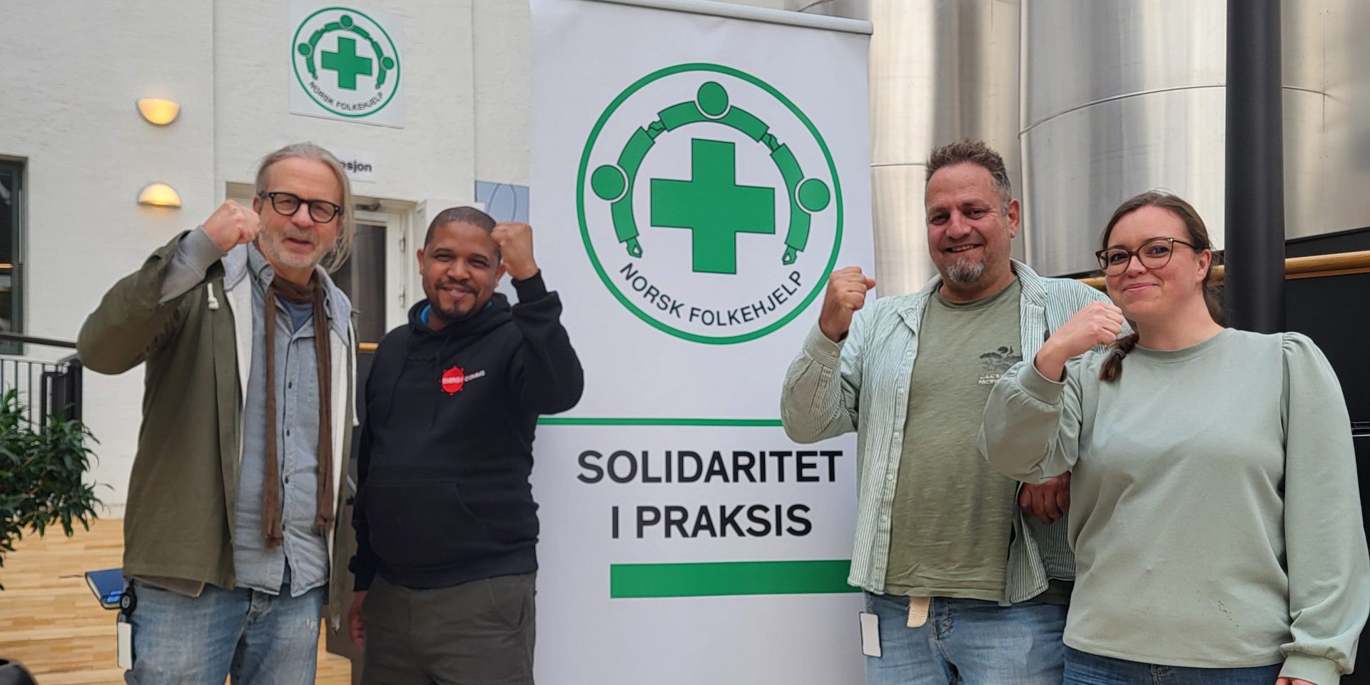 Arne Presterud, Thapelo Mohapi, Frank Toks og Naemy Trandum Aasen står foran en plakat med Norsk Folkehjelps logo og teksten "Solidaritet i praksis," mens de løfter knyttede never som symbol på solidaritet.