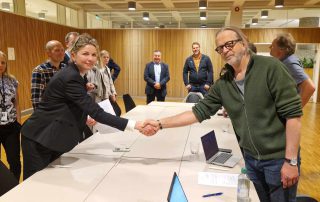 Nestleder Arne Presterud og Iselin Bauer Seeberg fra NHO Byggenæringen håndhilser over et møtebord i en konferansesal, mens flere personer står i bakgrunnen og ser på. På bordet ligger det dokumenter og bærbare datamaskiner.