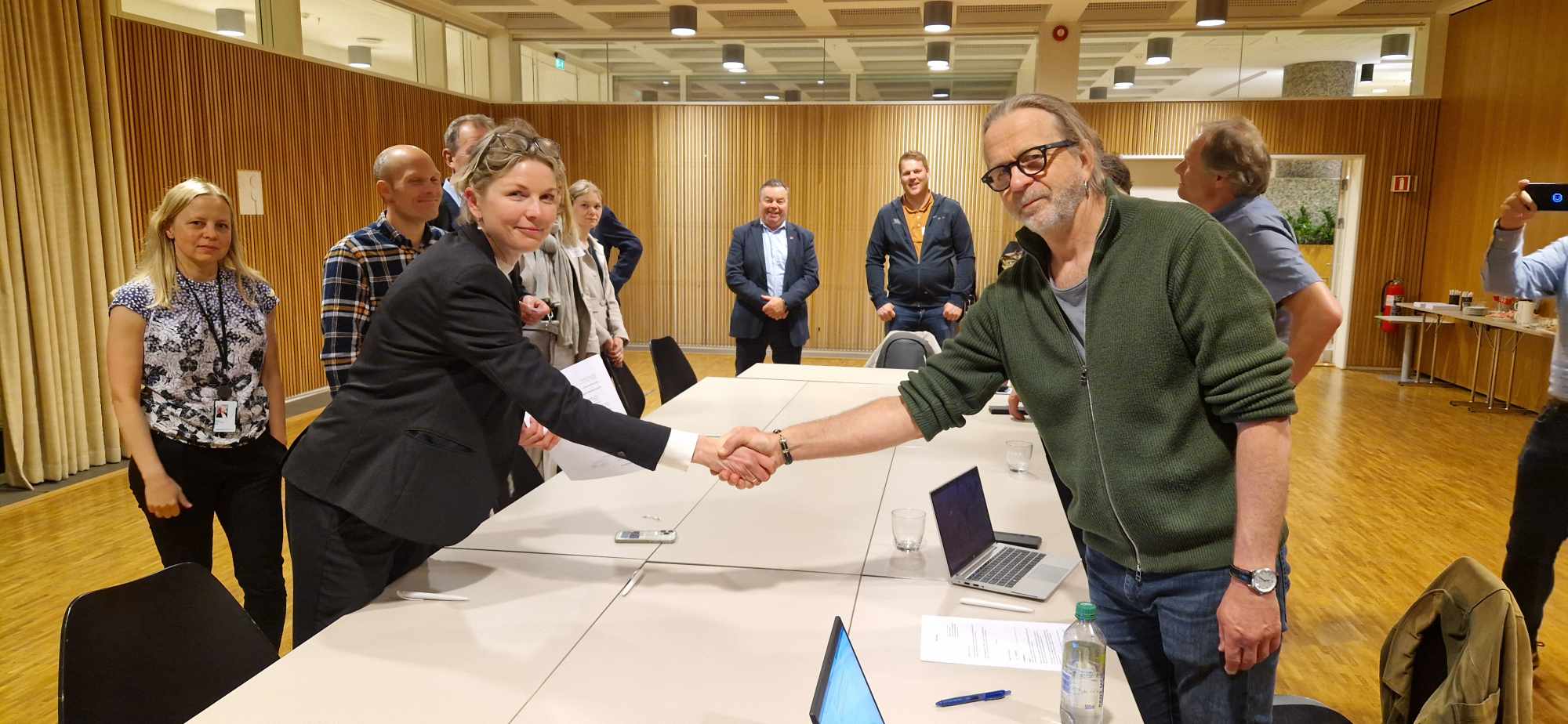 Nestleder Arne Presterud og Iselin Bauer Seeberg fra NHO Byggenæringen håndhilser over et møtebord i en konferansesal, mens flere personer står i bakgrunnen og ser på. På bordet ligger det dokumenter og bærbare datamaskiner.
