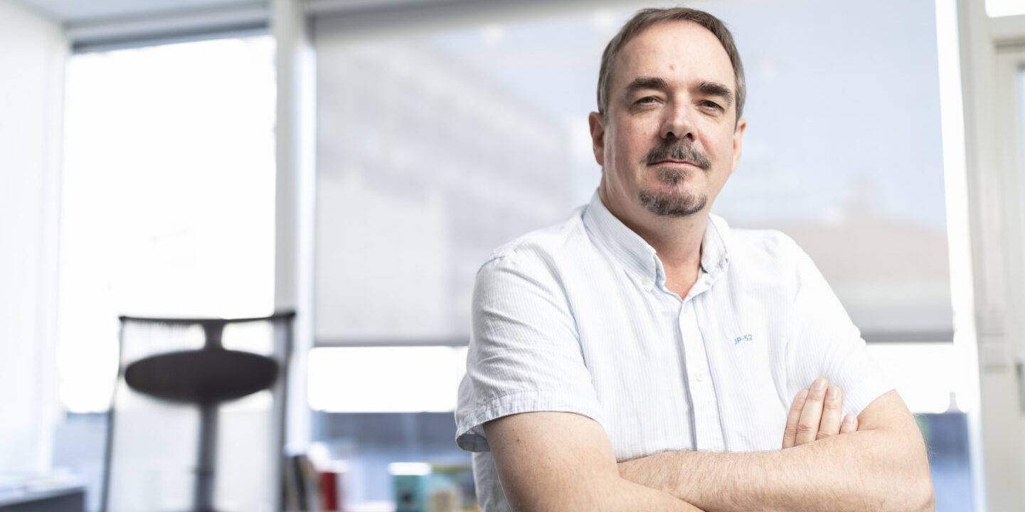 Portrett av Brede Edvardsen, leder i Norsk Arbeidsmandsforbund, som står med armene i kors i et lyst kontorlokale. Han ser fornøyd ut. Bakgrunnen viser et vindu med gardiner og en kontorstol.
