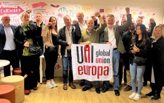 En gruppe personer står samlet foran en vegg med illustrasjoner, holder opp nevene i en gest av solidaritet, og holder et banner med teksten "UNI Global Union Europa".