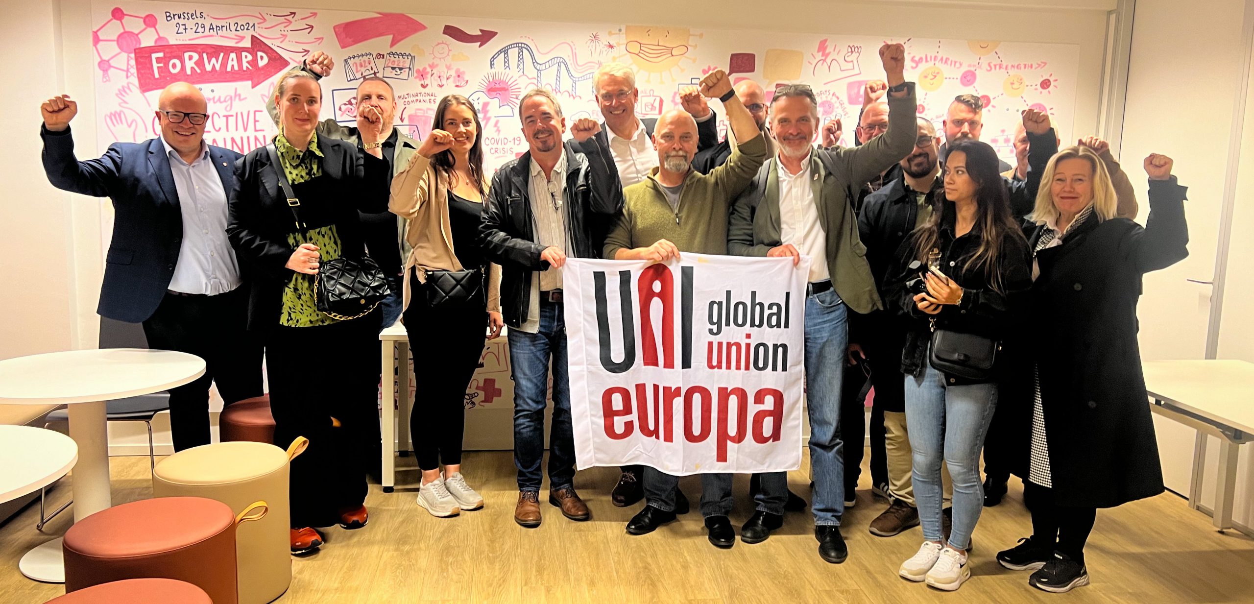 En gruppe personer står samlet foran en vegg med illustrasjoner, holder opp nevene i en gest av solidaritet, og holder et banner med teksten "UNI Global Union Europa".