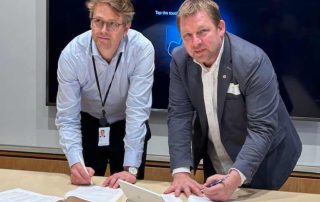 To dresskledde menn står ved et bord og signerer dokumenter. Begge ser opp mot kameraet.