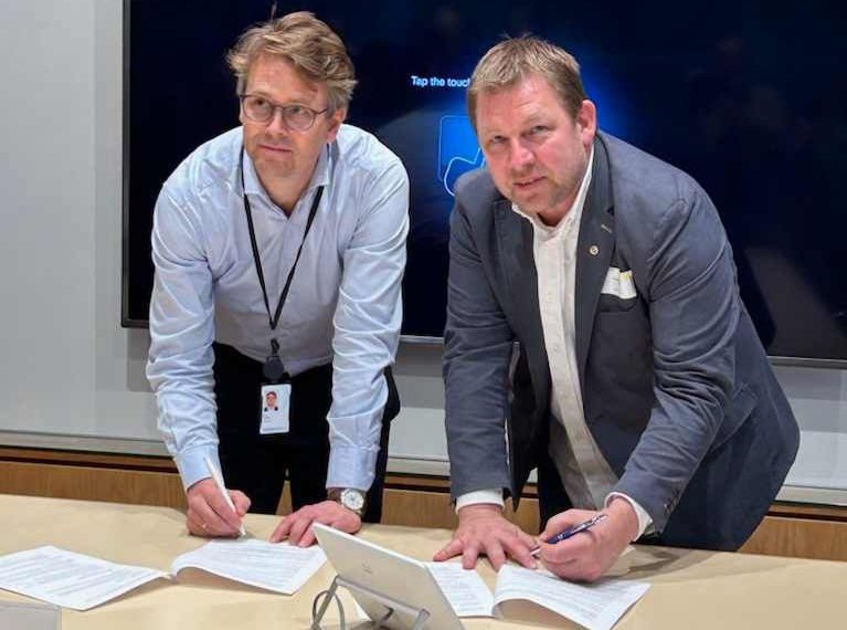 To dresskledde menn står ved et bord og signerer dokumenter. Begge ser opp mot kameraet.