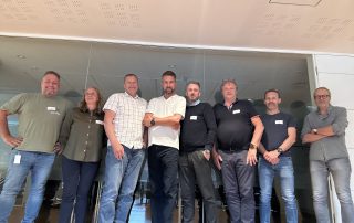 En gruppe mennesker fra forhandlingsdelegasjonen står samlet foran en glassvegg, smilende og side om side.